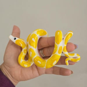 May include: A small, coiled snake figurine featuring a bright yellow and white patterned design. The snake has a smooth, glossy finish with a distinct head, a single visible black eye, and a curved body that forms a loop in the center.