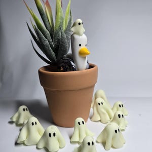 Puede incluir: Una maceta de terracota con una suculenta artificial y una figura de pato de cerámica con un fantasma encima. Varias pequeñas figuras de fantasmas blanquecinos están esparcidas alrededor de la maceta, creando una exhibición caprichosa con temática de Halloween.