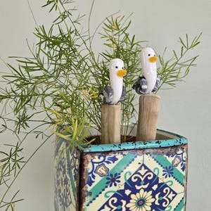 May include: A square, decorative planter with a blue, green, and cream geometric pattern. Two ceramic bird figurines with white bodies, gray wings, and yellow beaks sit atop wooden posts. The planter holds a green, leafy plant.
