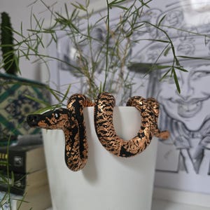 May include: A black and copper snake sculpture coiled around a white ceramic pot. The snake has a textured pattern and is among green plants. The pot is on a white surface, with books and artwork in the background.