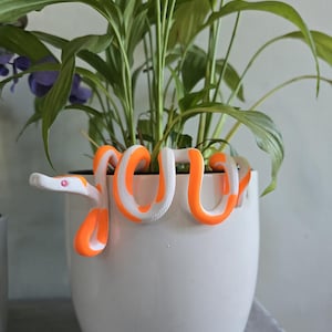 Könnte beinhalten: Ein weißer Keramik-Pflanzentopf mit einer dekorativen orange-weiß gestreiften Schlange, die sich um den Rand windet. Der Topf enthält eine grüne Blattpflanze mit lila Blüten. Die Schlange hat ein rosa Auge.