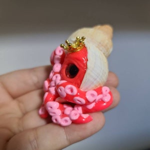 May include: A pink and red octopus figurine with a gold crown, holding a seashell. The octopus has a large eye and is made of polymer clay.