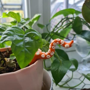 May include: A small orange and white octopus figurine is placed in a pink pot with a green plant. The octopus is curled up and has a white dot pattern.