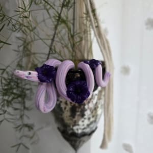 May include: A decorative hanging planter with a distinctive design. It features a light purple and white striped snake coiled around a pot filled with dark purple flowers. The planter is suspended by a macrame hanger, with green foliage in the pot.