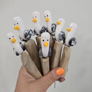 Può includere: Una collezione di picchetti da giardino a forma di gabbiano fatti a mano. Ogni picchetto presenta un gabbiano bianco con becco giallo e ali a fantasia bianca e nera. I picchetti sono attaccati a tasselli di legno.