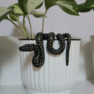 Könnte beinhalten: Ein weißer, gerippter Keramik-Blumentopf mit schwarzem Rand, der eine Pothos-Pflanze enthält. Eine schwarze, gefleckte, schlangenförmige Dekoration ist über den Rand drapiert. Die Schlange hat gelbe Punkte und windet sich um die Pflanze.