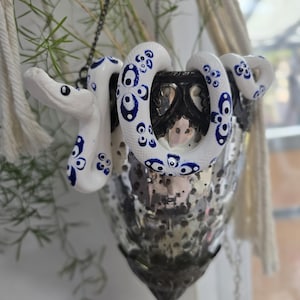 Op de afbeelding: Een decoratieve hangende glazen kandelaar met een witte en blauwe keramische slang eromheen gewikkeld. De slang heeft blauwe cirkelvormige patronen en lijkt de kandelaar te bewaken. De kandelaar heeft een zilveren afwerking.