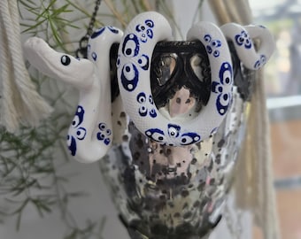 May include: A decorative hanging glass candle holder with a white and blue ceramic snake wrapped around it. The snake has blue circular patterns and is positioned to appear as if it is guarding the candle holder. The candle holder has a silver finish.