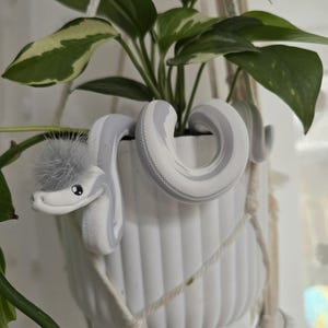 Könnte beinhalten: Ein weißer Keramik-Pflanztopf mit einer grau-weißen Schlangen-Dekoration, die sich darum windet. Der Topf wird von einem Makramee-Aufhänger gehalten und enthält eine grüne Pflanze mit panaschierten Blättern. Die Schlange hat einen flauschigen grauen Kopf.