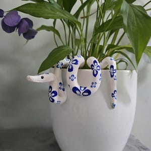 Peut inclure: Un pot de fleurs en céramique blanche avec un motif de serpent décoratif peint à la main en bleu. Le serpent est enroulé autour du pot, qui contient une plante verte avec de grandes feuilles et des fleurs violettes.