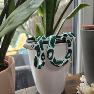 May include: A white ceramic plant pot accessory shaped like a snake with a glossy finish. The snake features a pattern of dark green spots and patches along its body, with a pointed head and visible eyes, designed to hook over the rim of a planter.
