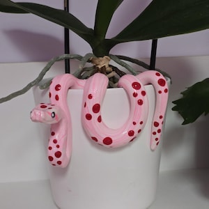 May include: A white plant pot decorated with a pink snake with red spots. The snake coils around the pot, which contains a green plant with long leaves. The pot is made of ceramic.