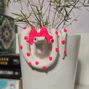 Könnte beinhalten: Ein weißer Keramik-Pflanztopf mit einem Schlangenmuster. Die Schlange ist weiß mit pinkfarbenen Herzakzenten und einer leuchtend pinkfarbenen Schleife. Der Topf enthält grüne Pflanzen.