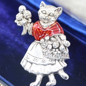 May include: A silver cat figurine wearing a red and white dress and carrying a basket of flowers. The figurine is in a blue velvet box.