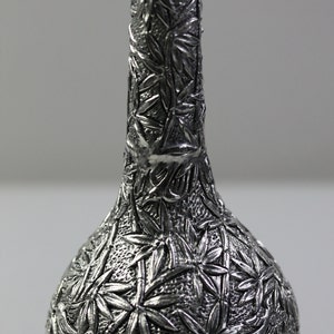 May include: A silver decorative bottle with an intricate design of leaves and flowers. The bottle has a round body and a long, slender neck.
