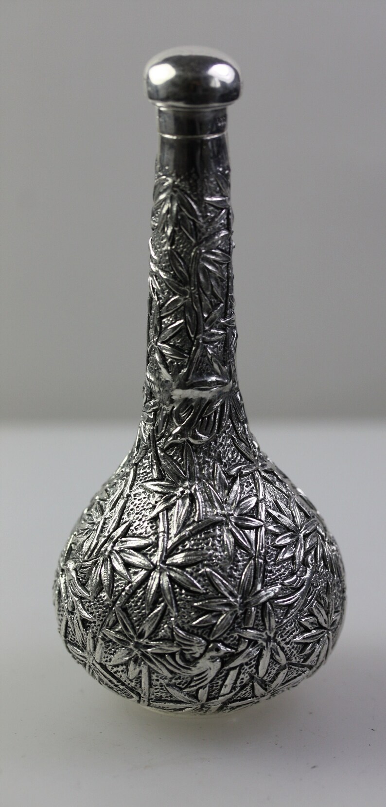 May include: A silver decorative bottle with an intricate design of leaves and flowers. The bottle has a round body and a long, slender neck.