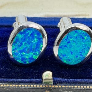 May include: A pair of silver cufflinks with oval-shaped, blue-green opal stones. The cufflinks are set in a silver frame and are displayed in a blue velvet box. The box has the text "GASS & Co. 138, REGENT STREET, LONDON."