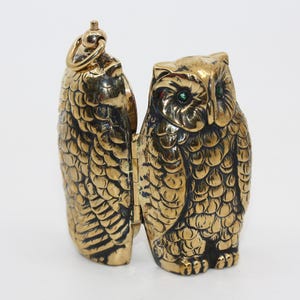 Könnte beinhalten: Eine goldfarbene, eulenformige Schmuckschatulle, die geöffnet ist, um ihr Inneres zu zeigen. Die Eule hat detaillierte Federmuster und grüne Augen. Die Box ist aus Metall und hat ein Scharnierdesign.