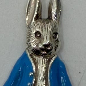 May include: A silver rabbit figurine with a blue jacket. The rabbit has detailed fur and long ears. The jacket is a bright blue with a glossy finish and small silver buttons. A charming decorative item.