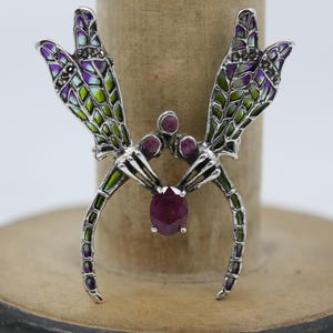 Puede incluir: Broche de libélula plateada con detalles intrincados. Las alas presentan un patrón de mosaico en tonos morados, verdes y blancos. El cuerpo del broche está adornado con una gran gema de rubí ovalada y pequeños acentos de rubí.