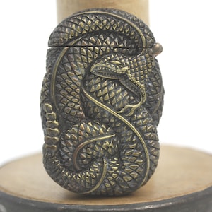 May include: A bronze-colored, intricately carved box featuring a coiled snake design. The snake's scales and details are finely rendered, with a hinged lid and a small, round clasp. The box is set on a wooden base.