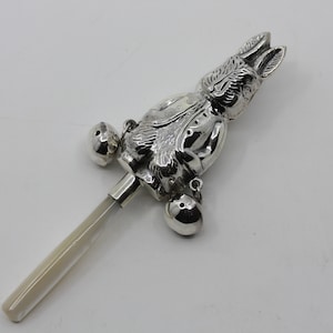 May include: A silver baby rattle in the shape of a bunny, featuring two bells and a handle. The bunny has detailed features, including ears and facial features. The handle is a light cream colour.