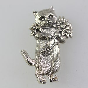 Op de afbeelding: Zilverkleurige kattenbroche. De kat staat rechtop en houdt een boeket bloemen vast. De broche heeft een gedetailleerd ontwerp, met zichtbare vacht en gezichtskenmerken. De staart van de kat is gekruld.