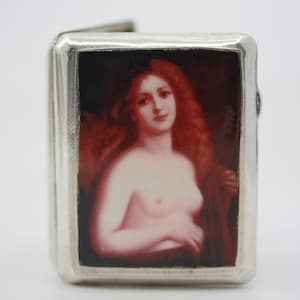 May include: Silver cigarette case with a rectangular portrait of a nude woman with red hair. The portrait is framed by a dark red background. The case has a hinged lid and a silver border.