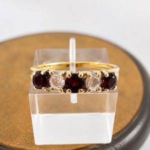 May include: A gold ring with five gemstones: alternating dark red and clear stones. The ring is presented on a clear stand. The gemstones are round cut and set in a gold band. The ring is displayed against a neutral background.