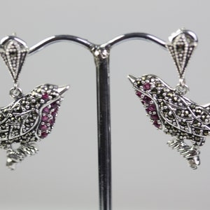May include: A pair of silver bird earrings. Each earring features a bird design with a body of small, dark stones and a cluster of red stones. The earrings hang from a decorative silver element.