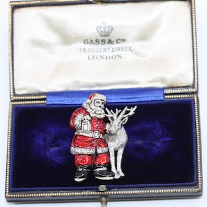 May include: A silver-toned brooch depicting a Santa Claus figure wearing a red and white suit, holding a lantern, and a reindeer by his side. The brooch is presented in a blue velvet box with gold trim and the inscription "GASS & Co. 138. REGENT STREET. LONDON."