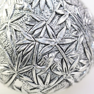 May include: A silver ornament with a detailed design of leaves and a bird. The ornament is round and has a smooth, shiny surface.