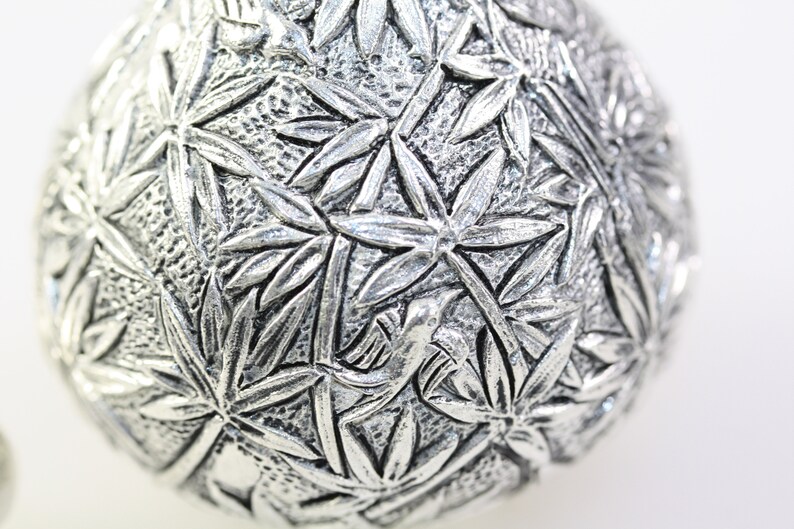 May include: A silver ornament with a detailed design of leaves and a bird. The ornament is round and has a smooth, shiny surface.