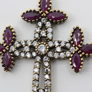 Puede incluir: Un colgante de cruz de plata con un diseño detallado que presenta una piedra central y piedras más pequeñas alrededor de los bordes. La cruz está adornada con piedras preciosas púrpuras.