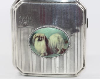 May include: Silver compact with a raised monogram "H.H." and a round enamel portrait of a Pekingese dog on the front.