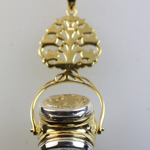 Könnte beinhalten: Ein Anhänger aus Gold und Silber mit einem Lebensbaum-Design und einem runden, klappbaren Behälter. Der Behälter hat eine silberne Oberseite und eine goldene Unterseite.