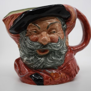 Op de afbeelding: Een keramische kan met een handvat, met een gedetailleerd gezicht van een man met een lange grijze baard en snor. De man draagt een zwarte hoed en een rode jas. De kan is beschilderd met tinten bruin, rood en grijs.