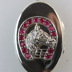 May include: Silver oval pendant featuring a horse head and horseshoe design. The horse head is centrally placed, encircled by a horseshoe shape set with red gemstones. The pendant has a bail for a chain.
