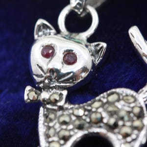 Può includere: Ciondolo a forma di gatto in argento con occhi di gemme rosse e un piccolo papillon. Il corpo del gatto è incrostato di piccole pietre scintillanti. Il ciondolo è sospeso da un anello di catena d'argento.