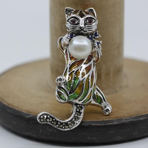Può includere: Spilla a forma di gatto argentato che tiene una grande perla. Il gatto ha occhi di pietre preziose rosse e blu e un design smaltato colorato sul corpo. La coda è decorata con piccole pietre scure.