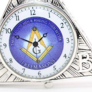 May include: Silver-toned triangular clock with a white face and blue center featuring Masonic symbols and the text "TAKING GOOD MEN & MAKING THEM BETTER" and "FREEMASONS". The clock has a classic design.