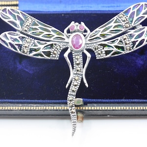 Puede incluir: Broche de plata con forma de libélula con alas iridiscentes, detalles de marcasita y un rubí ovalado central. El broche se muestra en un joyero de terciopelo azul con ribete negro y dorado.