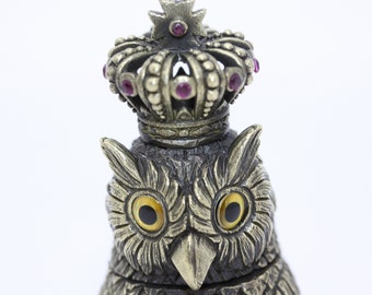May include: A silver owl figurine with a crown on its head. The crown is decorated with pink jewels and the owl has yellow eyes.