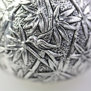 May include: A silver ornament with a detailed design of bamboo leaves and a bird in flight.