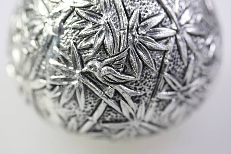 May include: A silver ornament with a detailed design of bamboo leaves and a bird in flight.