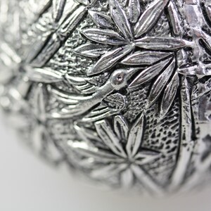 May include: A silver ornament with a detailed design of leaves and a bird. The ornament is round and has a textured surface.