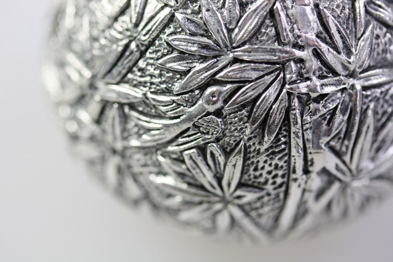 May include: A silver ornament with a detailed design of leaves and a bird. The ornament is round and has a textured surface.