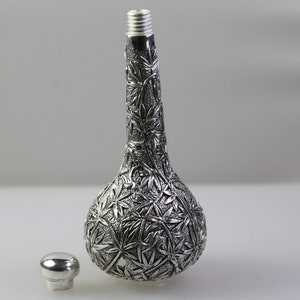 May include: A silver bottle with a detailed embossed design of leaves. The bottle has a round body and a long, thin neck. The bottle is sitting on a white surface.