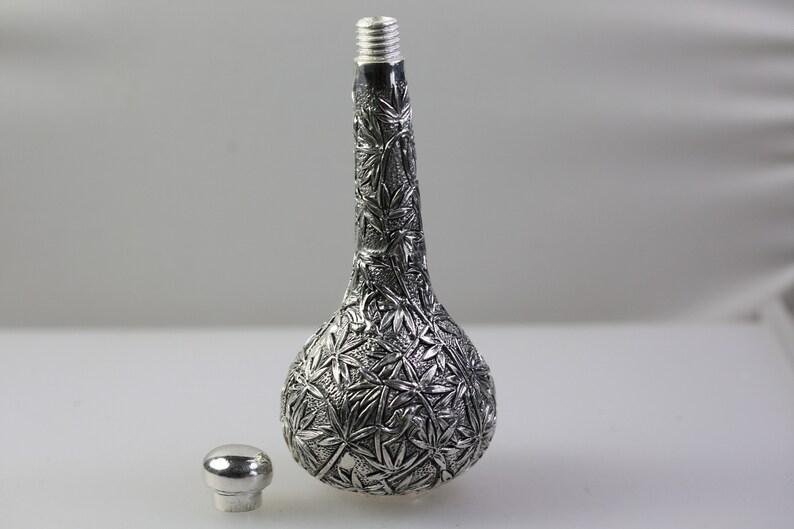 May include: A silver bottle with a detailed embossed design of leaves. The bottle has a round body and a long, thin neck. The bottle is sitting on a white surface.