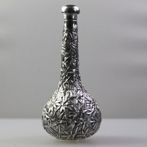 May include: A silver decorative bottle with an intricate floral design. The bottle has a round base and a long, slender neck.